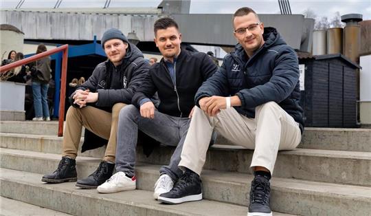 Drei Männer sitzen auf einer Treppe vor einer Brücke und tragen Freizeitkleidung mit Jacken und Sneakers