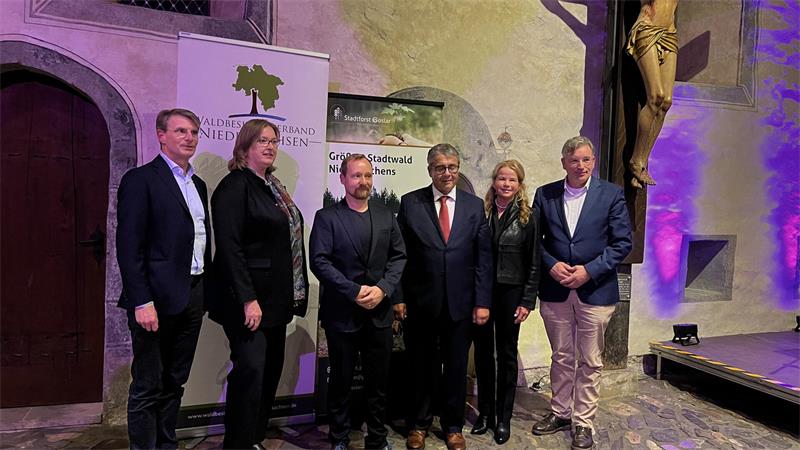 Sechs Personen stehen bei einer Veranstaltung in einem historischen Innenraum vor Bannern und einer beleuchteten Bühne. Im Hintergrund ist ein großes Kruzifix zu sehen.
