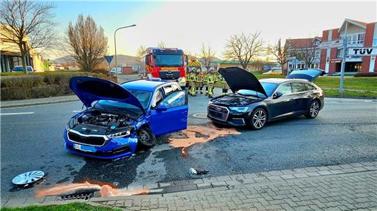 Zwei beschädigte Autos mit geöffneten Motorhauben und einer offenen Fahrertür auf einer Straße, im Hintergrund Feuerwehrleute und ein Feuerwehrauto