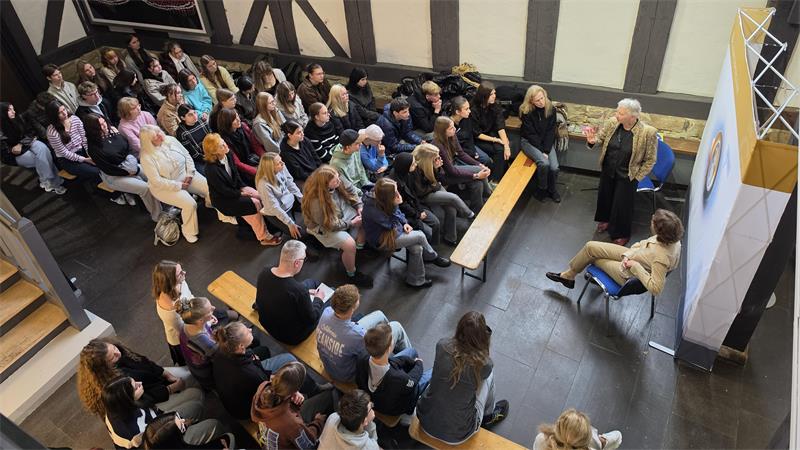 Blick von oben in einen Fachwerkraum, in dem viele Menschen auf Bänken sitzen und einer Frau zuhören, die vor ihnen steht.