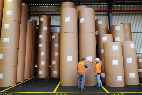 Zwei Arbeiter in einer Papierfabrik vor verpackten Papierrollen.