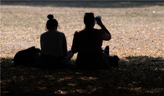 Zwei Frauen sitzen in einem Park im Schatten der Bäume, um sich vor der Hitze zu schützen.