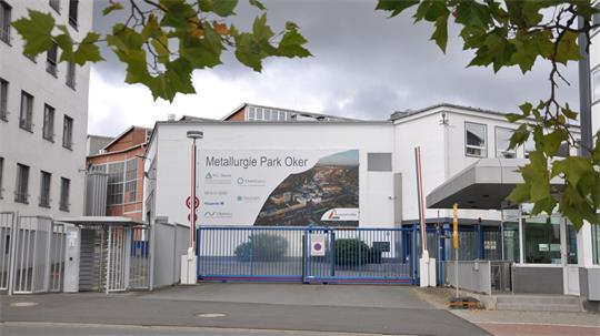 Eingangstor mit blauem Gitter vor einem weißen Gebäude und einem großen Werbebanner mit der Aufschrift 'Metallurgie Park Oker'