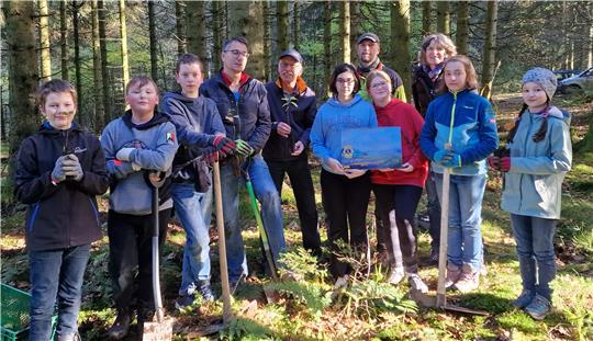 Kinder und Erwachsene stehen im Wald. Einige Kinder halten einen Spaten in der Hand, andere einen Buchen-Setzling.