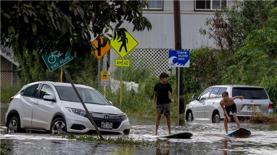 Zwei Jugendliche surfen in Waialua neben einem liegengebliebenen Fahrzeug im Hochwasser.