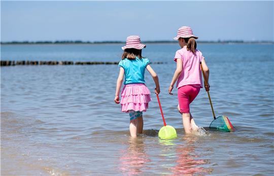 Zwei Mädchen spielen bei sonnigem Wetter mit ihren Keschern im flachen Wasser der Nordsee am Strand.