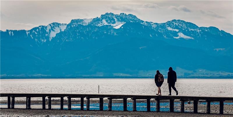 Wintereinbruch zum Wochenstart erwartet Zwei Männer gehen bei frühlingshaften Temperaturen vor der fast schneefreien Kulisse der Kampenwand in den Chiemgauer Bergen über einen Steg im Chiemsee.