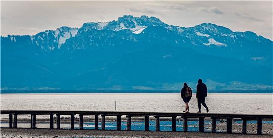 Zwei Männer gehen bei frühlingshaften Temperaturen vor der fast schneefreien Kulisse der Kampenwand in den Chiemgauer Bergen über einen Steg im Chiemsee.