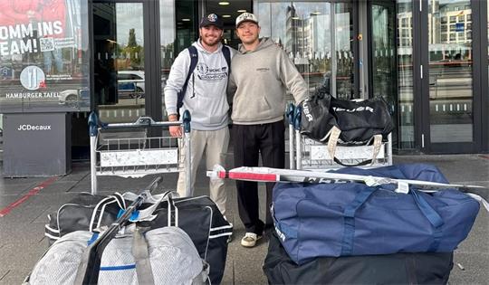 Zwei Männer mit Kappen stehen vor einem Flughafenterminal mit Gepäckwagen voller Taschen und Koffer