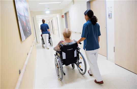 Zwei Pflegerinnen schieben Patienten mit Rollstühlen auf einem Flur in einem Krankenhaus. Foto: picture alliance/dpa | Daniel Bockwoldt