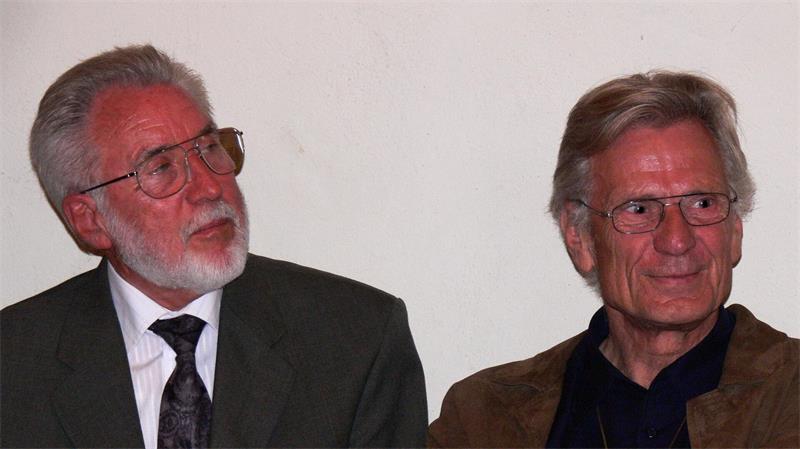 Zwei ältere Männer mit Brillen, der linke mit grauem Bart und Anzug, der rechte mit kurzem grauem Haar und dunklem Hemd, vor weißer Wand