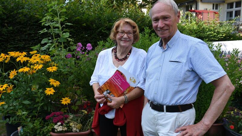 Eine Frau und ein Mann sehen im blühenden Garten. Die Frau hält ein Buch in der Hand.