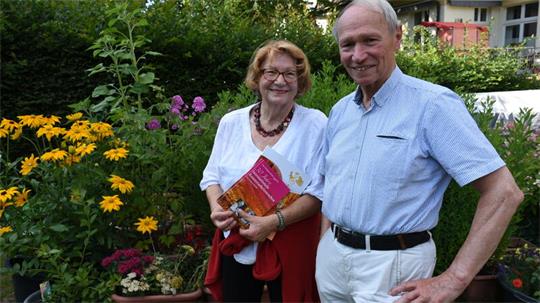Eine Frau und ein Mann sehen im blühenden Garten. Die Frau hält ein Buch in der Hand.