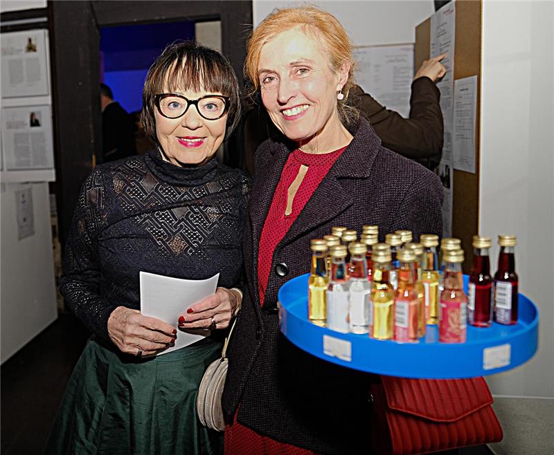 Eine Frau bietet lächelnd auf einem blauen Tablett kleine Schnapsfläschchen an, neben ihr lächelt eine Frau mit einem Papier in der Hand und schwarzer Brille in die Kamera.